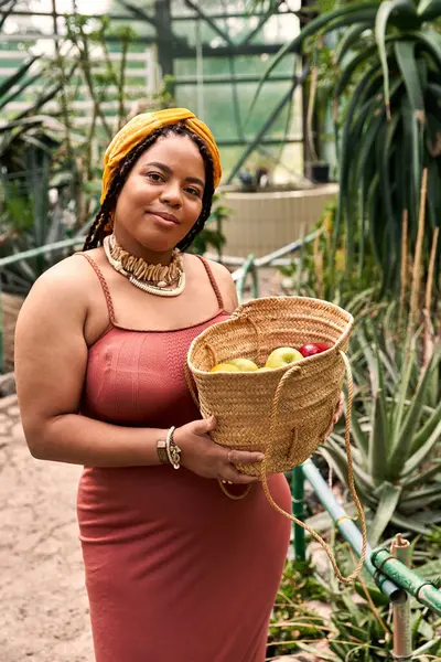 Örgülü bir kadın elinde meyve sepeti tutarken seranın güzelliğinden zevk alır..