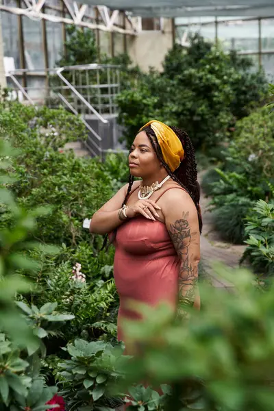 Saçları örgülü mutlu bir Afrikalı Amerikalı kadın dingin bir serada canlı bitkilerin arasında rahatlıyor..