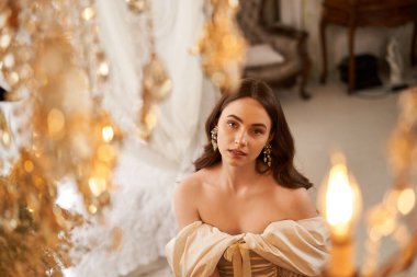 Zarif bir zarafete sahip genç bir kadın şık bir elbiseyle poz veriyor, etrafı lüks dekorlarla çevrili..