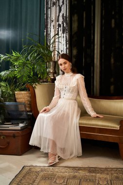 Dalgalı elbiseli genç bir kadın, yeşilliklerle çevrili eski bir kanepeye zarif bir şekilde oturur..