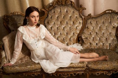 Genç bir kadın eski bir kanepeyi süslüyor, güzel bir elbise ve sakin bir ortam içinde zarafet yayıyor..