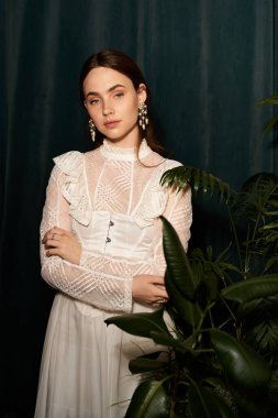 Güzel, genç bir kadın çiçeklerin arasında zarif duruyor. Kaliteli elbisesi zarafetle parıldıyor..