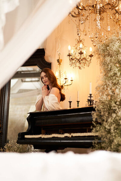 A graceful young woman in elegant attire stands by a piano, radiating timeless beauty.