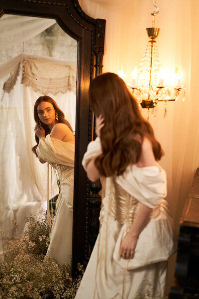 Красивая молодая женщина в потрясающем платье смотрит в винтажное зеркало, окруженное мягким декором.