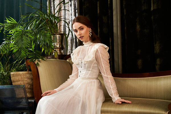 Красивая молодая женщина в винтажном платье элегантно сидит на диване в окружении пышной зелени.