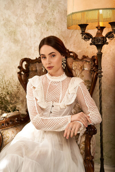 Молодая женщина в винтажном платье изящно сидит в украшенном кресле, излучая элегантность.