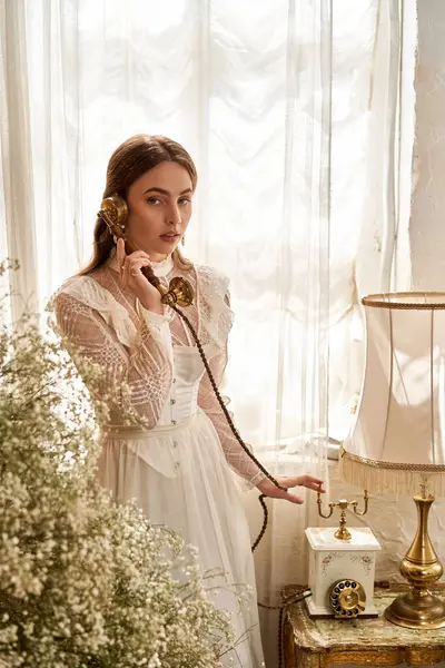 Klasik elbiseli genç bir kadın eski bir telefonu tutuyor, doğal ışıkla yıkanmış..