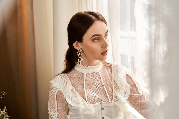 A beautiful young woman dressed in vintage attire gazes thoughtfully through a light-filled window.