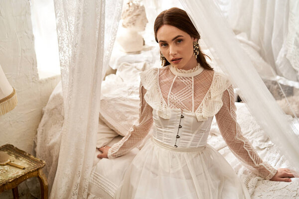 A beautiful young woman poses gracefully in an elegant vintage dress surrounded by soft fabrics.