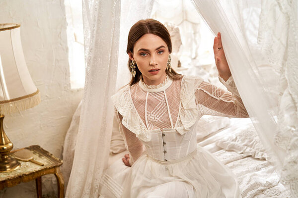 A beautiful young woman in a vintage dress sits elegantly, surrounded by soft, flowing fabrics.