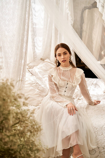 A young woman in a vintage dress sits gracefully amid soft fabrics and delicate decor.