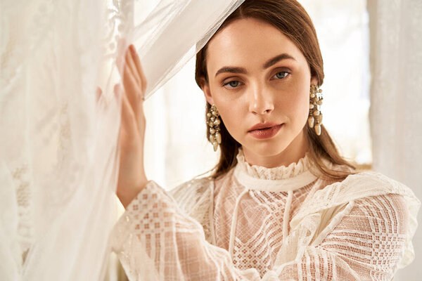 A graceful young woman in a vintage dress stands by flowing curtains, radiating beauty.