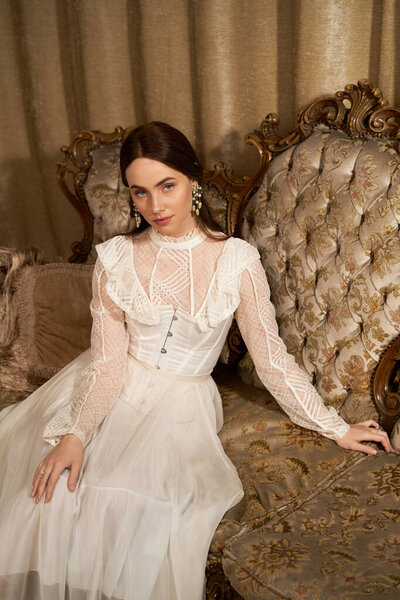 Красивая молодая женщина в винтажном платье изящно сидит на украшенном диване, излучая элегантность.