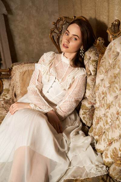 Молодая женщина в винтажном платье элегантно сидит на украшенном диване.