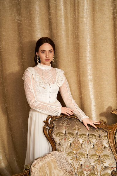 Молодая женщина в винтажном платье стоит у декоративного стула в элегантном декоре.