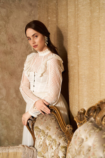 Молодая женщина в винтажном платье изящно прислоняется к украшенному стулу, источая элегантность.