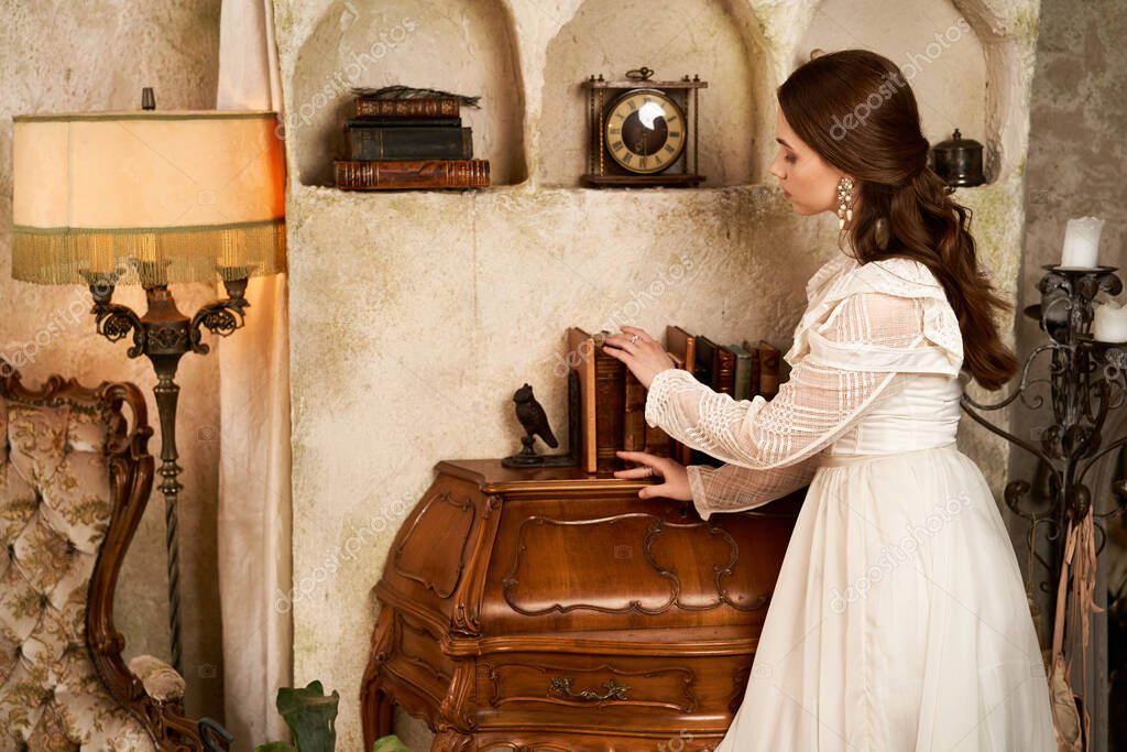 A young woman in a flowing dress gracefully arranges books in a vintage-inspired room.