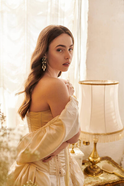 Молодая женщина в красивом платье задумчиво стоит у окна с мягким освещением, источая винтажное очарование.
