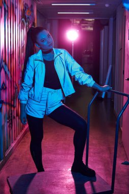 Kendine güvenen kadın, canlı neon ışıklarla aydınlatılmış bir şehir koridorunda modern moda sergiliyor..