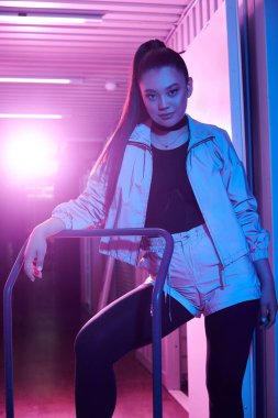 Kendine güvenen genç bir kadın dinamik neon ışıklı bir ortamda el arabasıyla zarif bir poz verir..