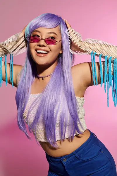 Mor saçlı ve son moda güneş gözlüklü şık bir kadın pembe bir fon karşısında mutlu bir şekilde parlıyor..