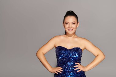 Güzel bir kadın kendine güvenerek duruyor, göz kamaştırıcı mavi elbisesiyle moda anlayışını sergiliyor..