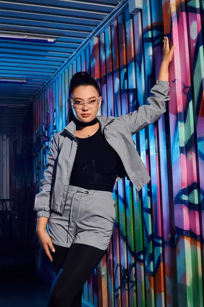 Kendine güvenen bir kadın renkli bir sokak resmine yaslanırken modern bir kıyafet sergiliyor..