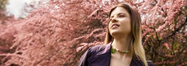 Renkli kolyeli genç bir kadın çiçek açan ağaçların arasında poz veriyor..