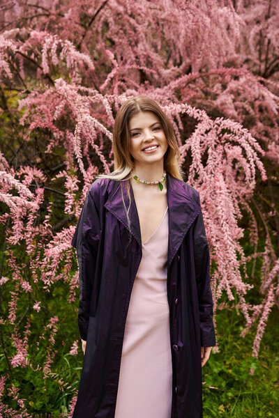 A young woman is standing and smiles gently, in front of a blossoming tree with pink flowers.