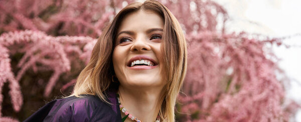 Smiling woman with long hair, pink blossoms, sunny day.