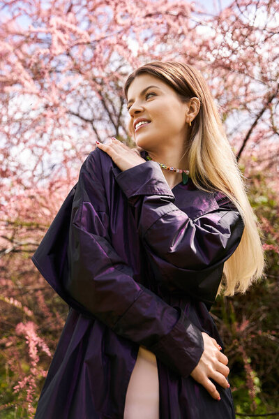 A pretty young woman in a jacket glances off towards the blossoms of a flowering tree in springtime.