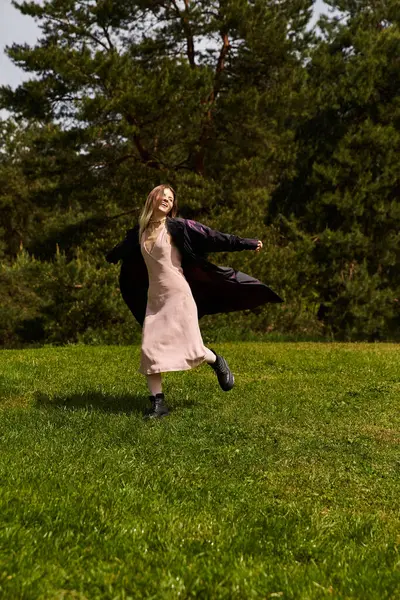 Açık renk saçlı bir kadın, elbise ve ceket giymiş, güneşli bir günde çimenli bir tarlada neşeyle kıvrılır..