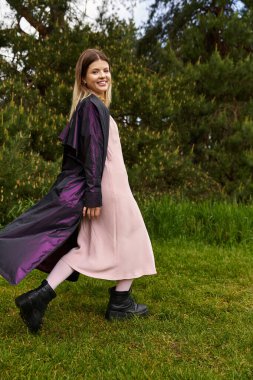 Gülümseyen kadın tarlada yürür, palto dalgalanır.