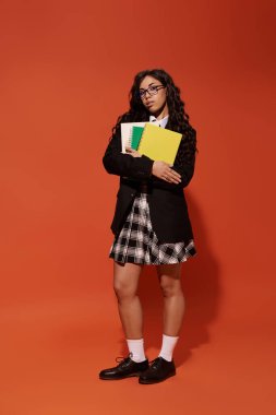 Güzel, genç bir kadın, şık bir okul kıyafeti içinde zarif bir şekilde poz verirken okul malzemelerini tutuyor..