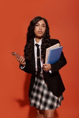 Güzel, genç bir kadın, elinde kitaplarla, elinde telefonla, kendinden emin bir şekilde ayakta duruyor..