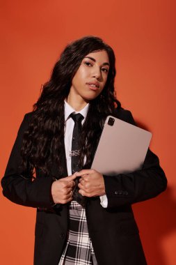 Genç bir kadın dikkat çekici bir turuncu arka plan karşısında duruyor, elinde bir tablet var..