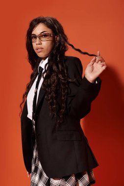 Kıvırcık saçlarından bir bukle tutan bir öğrenci resmi bir ceket ve desenli bir etekle poz veriyor..
