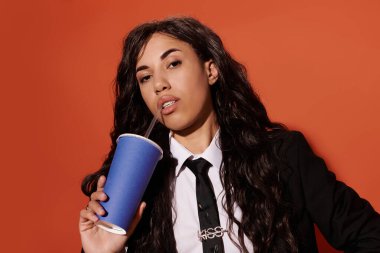 Genç bir kadın kendine güveni yayıyor, modaya uygun giyinmiş mavi bardaktan yudumlayarak, estetiğini gösteriyor..