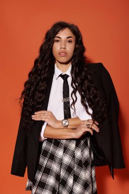 Genç bir kadın kendine güvenerek poz veriyor, turuncu arka planı olan şık bir öğrenci kıyafeti giyiyor..