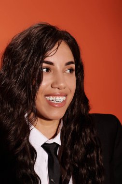 Diş teli takan genç bir kadın turuncu bir duvara karşı şık bir elbiseyle sıcacık gülümsüyor..