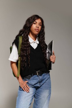 Kendine güvenen genç bir kadın, modern öğrenci estetiğini gösteren bir sırt çantası ve dizüstü bilgisayarla poz veriyor..