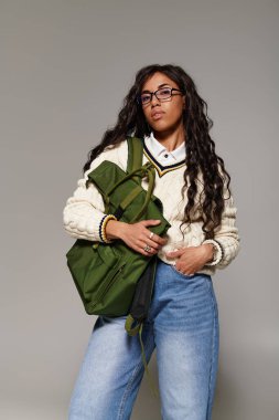 Güzel öğrenci rahat bir kazak ve kot pantolon içinde kendinden emin bir şekilde poz veriyor, gençlik enerjisi ve cazibesi yayıyor..