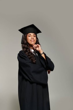 Mezuniyet kıyafetli kendine güvenen genç bir kadın gülümseyerek poz verir, gurur ve neşe saçar..