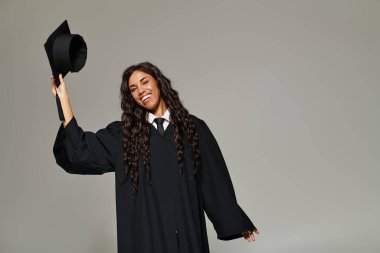 Genç bir kadın mezuniyet şapkasını parlak bir gülümsemeyle tutarak zaferin tadını çıkarıyor..