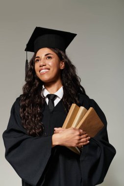 Mezuniyet elbisesi içindeki genç kadın kitaplarını tutarken gururla ışıldıyor, başarısını gösteriyor..