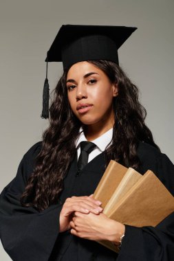 Parlak ışıklandırılmış arka plan, mezuniyet kıyafetiyle kitaplarını gururla tutan genç bir kadını çevreledi..