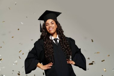 Mezuniyet şapkası ve cübbesi içinde genç bir kadın stüdyoda konfeti üzerine düşerken ışıl ışıl gülümsüyor..