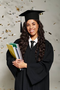 Önlüklü genç bir kadın konfeti düşerken elinde kitaplarla gülümsüyor..