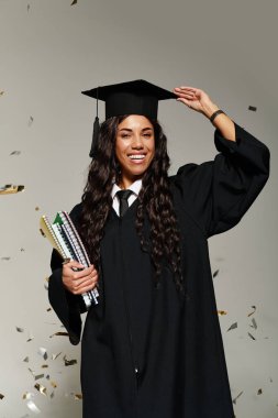 Mezuniyet şapkalı ve cüppeli genç kadın başarısını kitaplarla ve neşeyle kutluyor..
