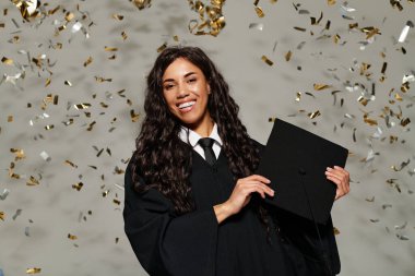 Genç bir kadın mezuniyet şapkasını sevinçle tutuyor, etrafında pırıl pırıl konfetiler var..